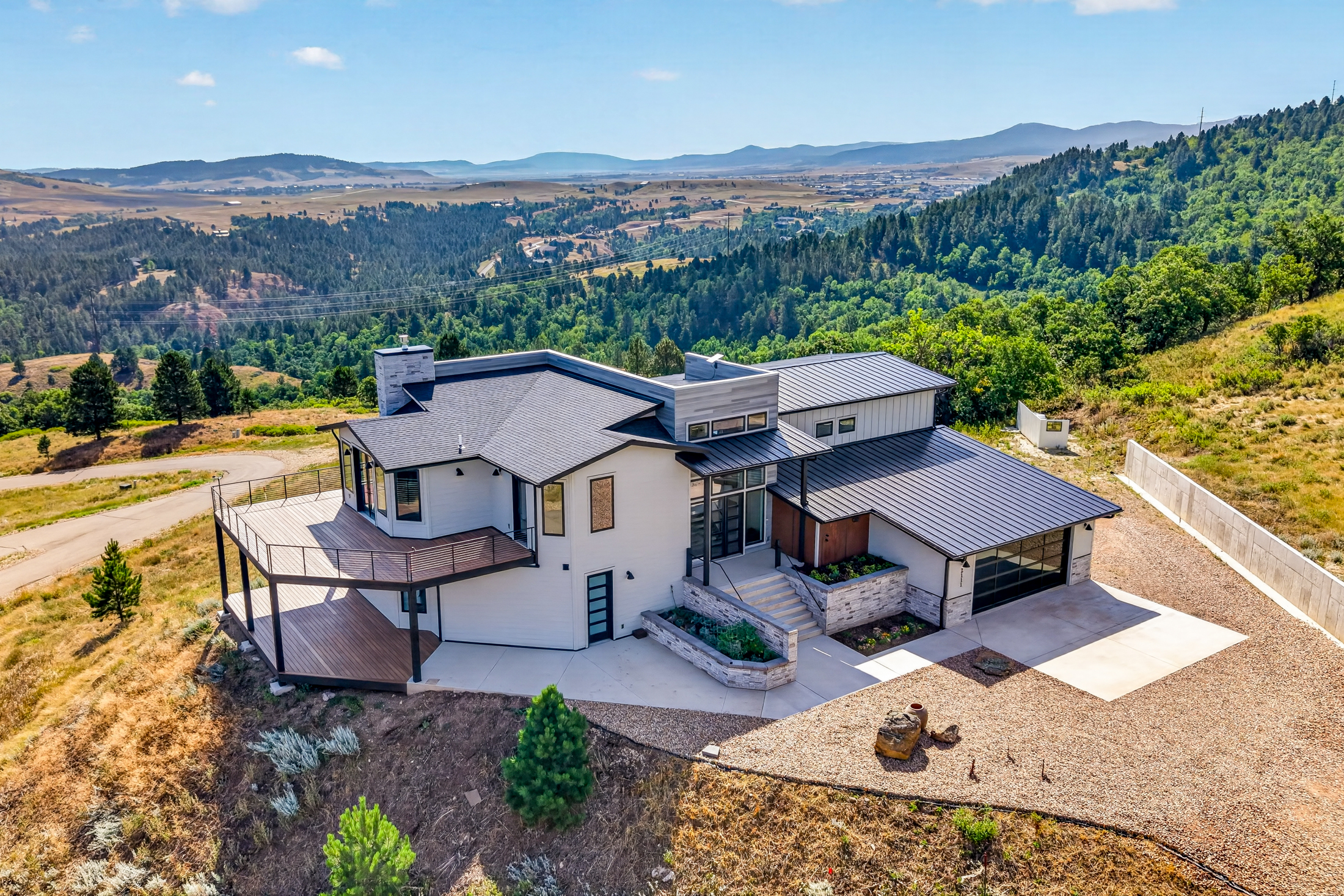 An aerial view shows 2111 Vantage Circle perched on a forested ridge above the Spearfish valley, with mountains layered in the distance.