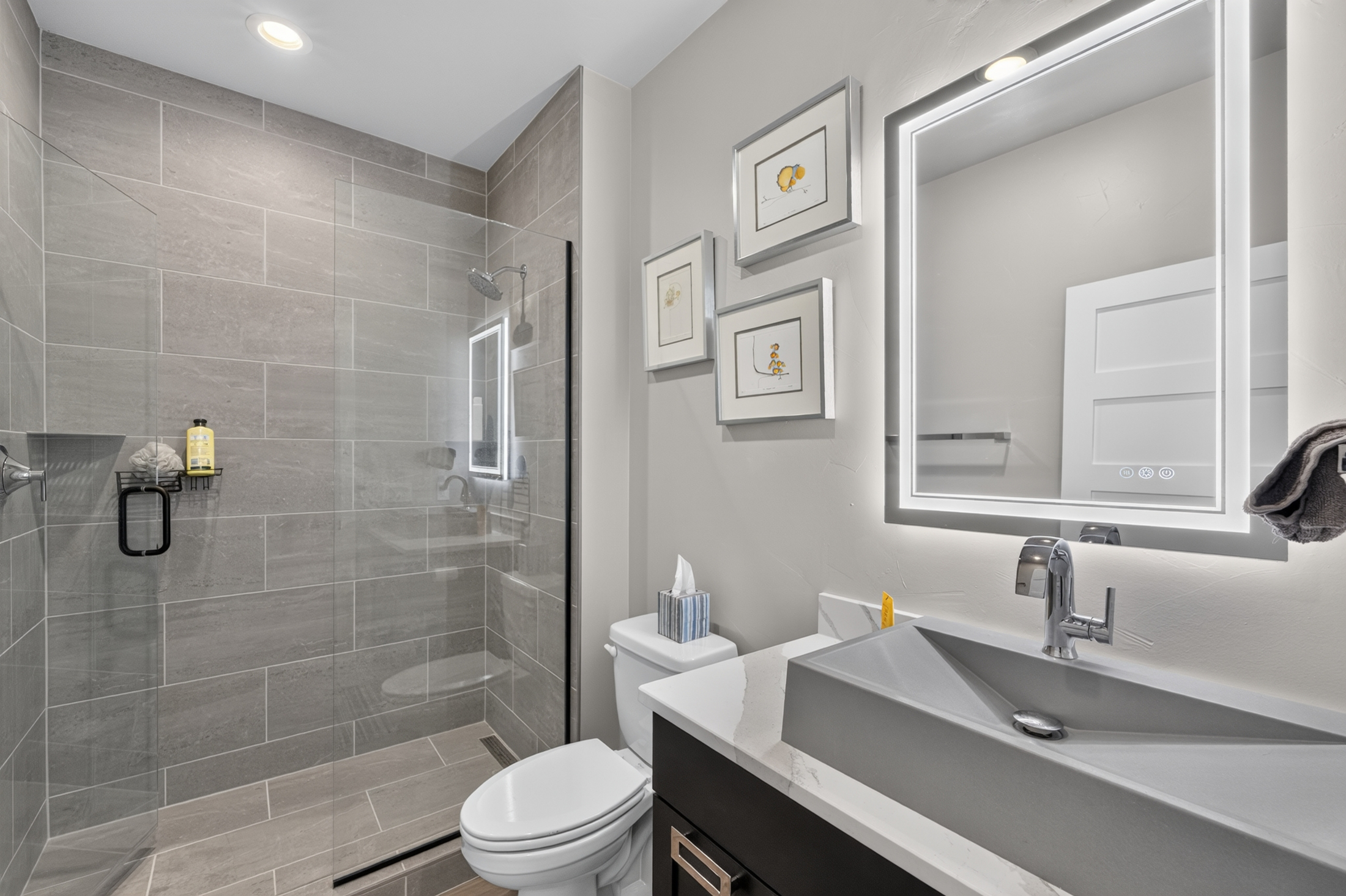 A guest bathroom pairs a row of small framed prints with a backlit mirror