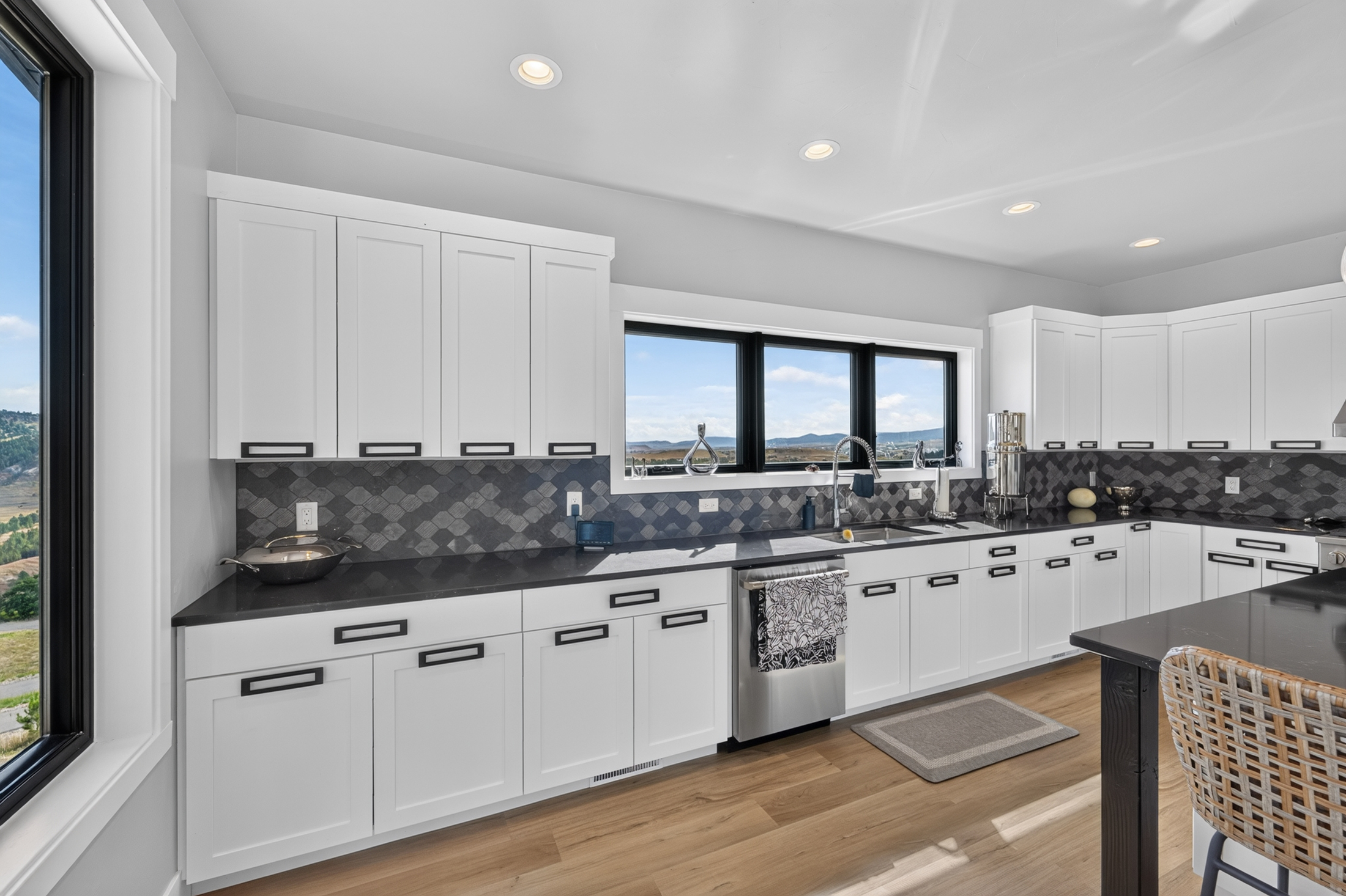 The kitchen sink sits beneath a wide window framing hills and open land, with a dishwasher and dark tile backsplash alongside.