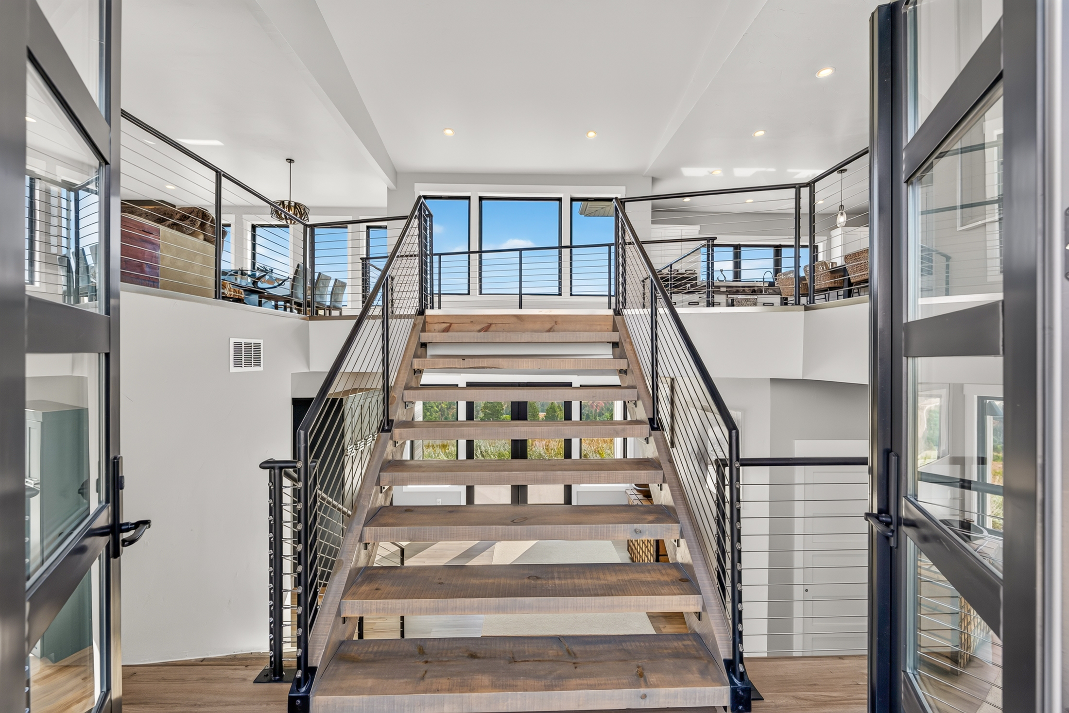 A wood-tread staircase with black cable railing rises to an open loft, framed by a tall window looking out over the hills.